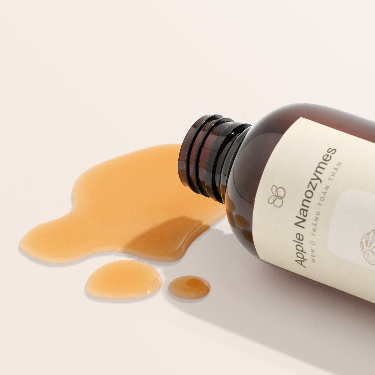 Ảnh tinh chất: chai toner nằm nghiêng, tinh chất vàng chảy ra tạo vũng trên nền be, chụp tại studio Juva Media