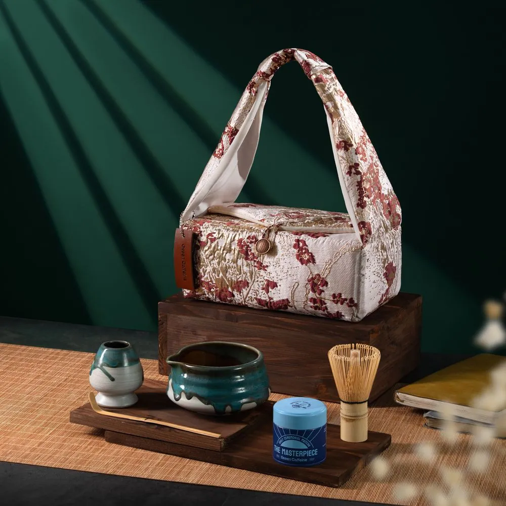 Túi vải hoa đặt trên khối gỗ cùng bộ pha matcha gồm bát, chổi tre và hũ bột trên chiếu tre, nền xanh rêu
